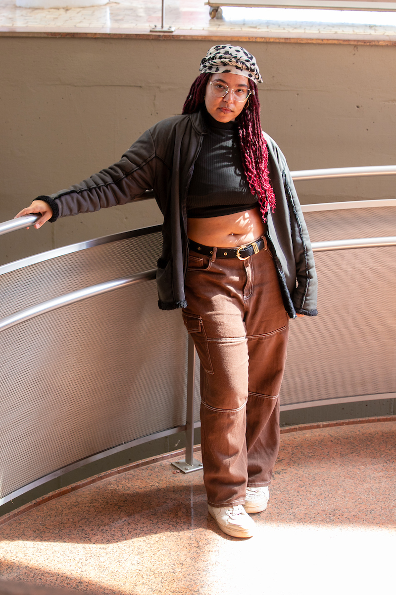 Garota posa para a foto. Cobrindo sua cabeça com um lenço, a jovem mantém a frente seu cabelo ruivo sobre seu casaco cinza e camisa cropped preta. Na inferior ela combina um cinto preto com detalhe dourado com sua calça marrom e tênis completamente branco.