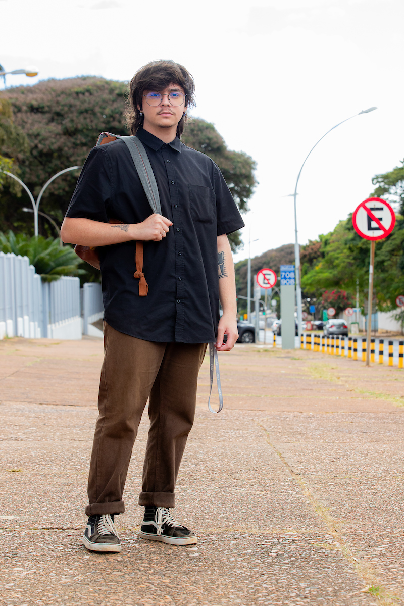 Rapaz posa para a foto. Apoiando sua mocilha cinza com a alça direita, o jovem veste uma camisa de botão preta combinando com uma calça marrom com a barra dobrada na altura do tornozelo e tênis cano médio na cor preta com detalhes em branco.