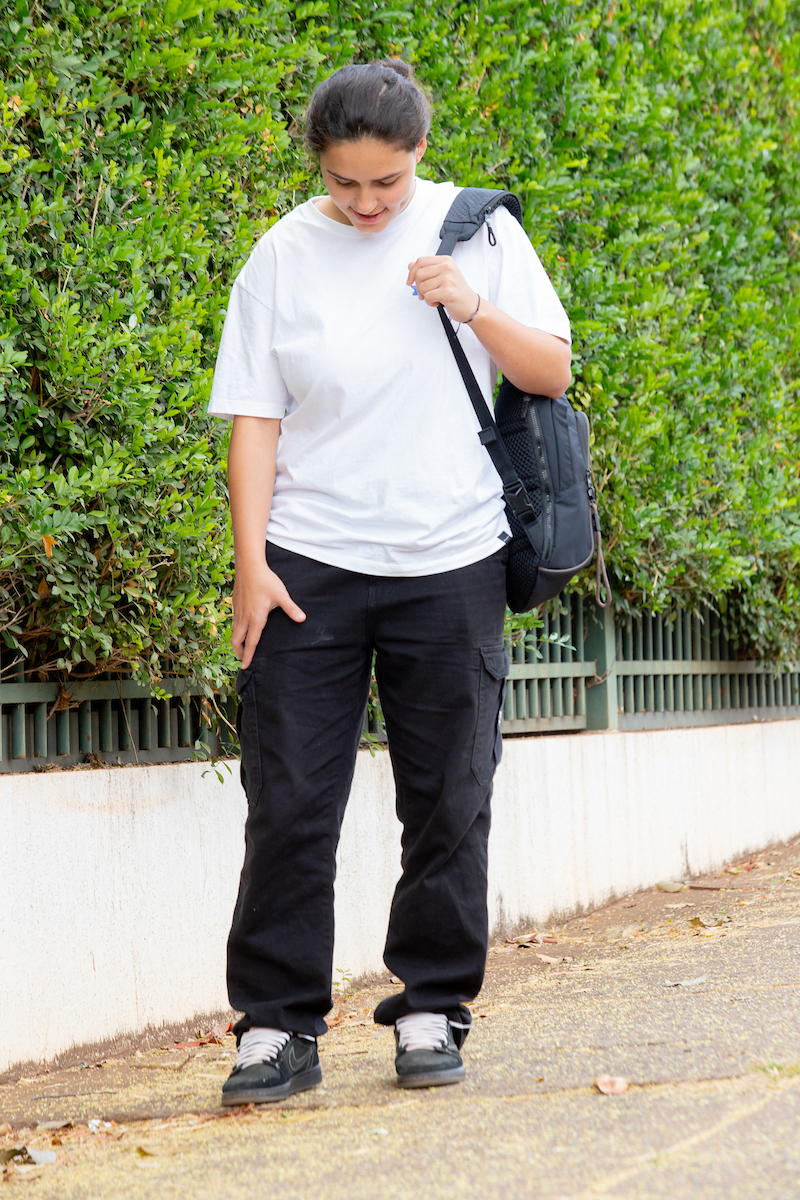 Moça posa para a foto. De camisa branca e cabelo preso, ela segura su mochila com o ombro esquerdo enquanto exibe sua calaça e tênis pretos, com detalhe do cadarço branco.