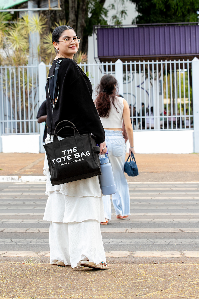 Mulher posa para foto em faixa de pedestres. com seu casaco preto vazado nas costas ela exibe sua bolsa também preta com estampa branca junto a sua calça branca com cortes sobrepostos.