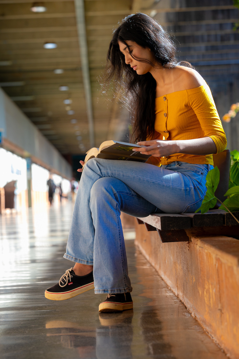 Garota posa para a foto. Com sua camisa de manga longa dobrada ao cotovelo, ela apoia seu livro em suas pernas vestindo calça jeans azul e um tênis cano baixo em azul, branco e vermelho.