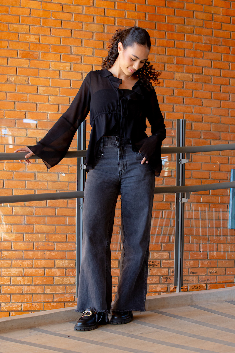 Garota posa para a foto. Com seu cabelo cacheado preso para trás, ela se apoia em um corrimão com sua camisa de renda preta e jeans escuro enquanto, combinando com seu sapato preto com detalhe dourado.