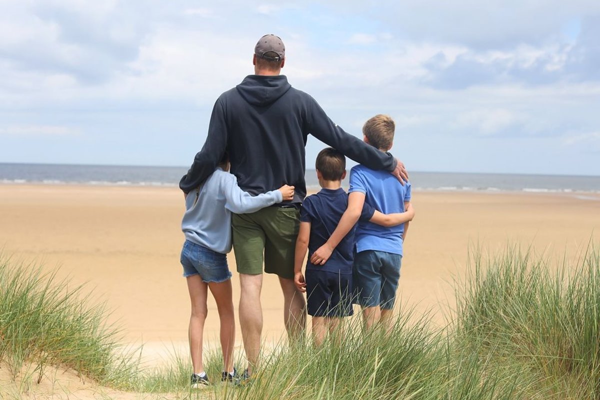 Foto colorida, William abraçado com os três filhos. Todos estão de costas olhando para o mar da areia da praia