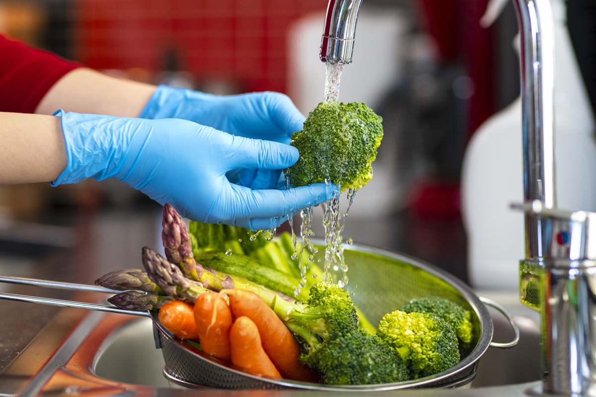 Imagem mostra as mãos de uma mulher usando luvas lavando alimentos em baixo de uma torneira - Metrópoles