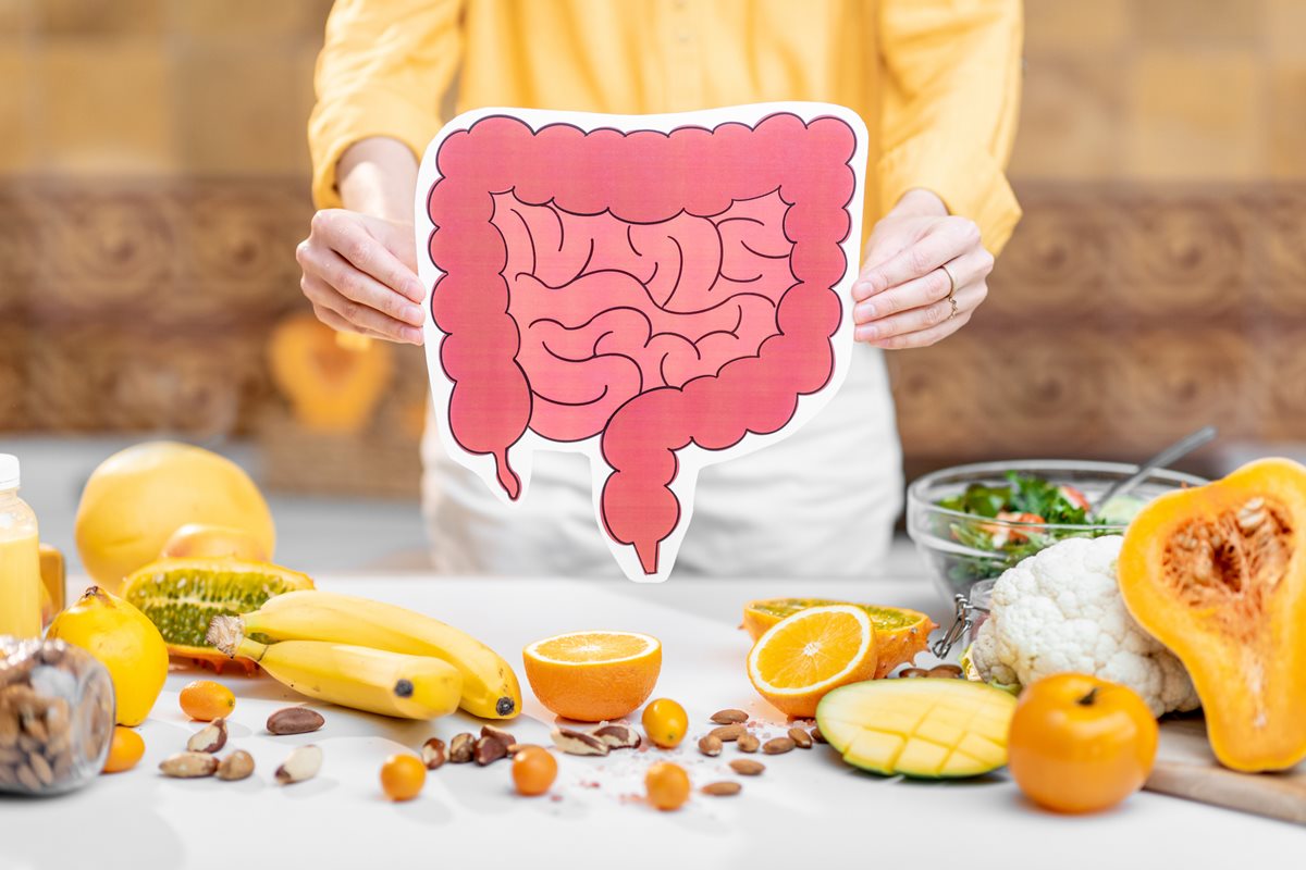 Foto colorida de mulher segurando o desenho de um intestino. Ela está próximo de uma mesa com frutas - Metrópoles