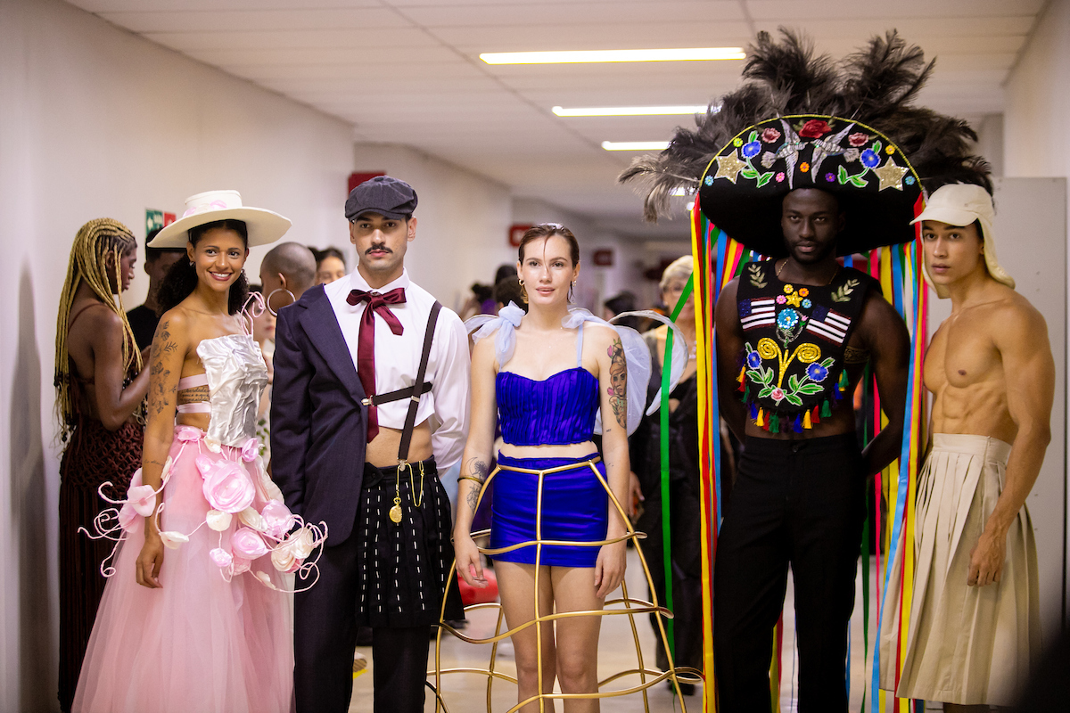 Cinco modelos posam para a foto. Na ponta esquerda, um jovem posa com vestido rosa e acessórios em branco. Ao seu lado, um rapaz posa com um terno dividido ao meio com uma meia saia apoiada por um suspensório. No centro da imagem, uma mulher com um conjunto azul com saia e camiseta posa com um acessório dourado. No seu lado direito, um homem aparece caracterizado como o Boi-Bumbá e à direita da imagem um rapaz posa sem camisa, com boné e saia bege.