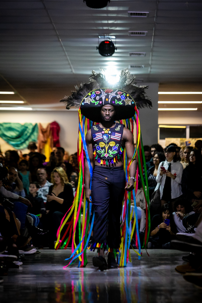 Homem caminha em passarela caracterizado como o Boi-Bumbá. Nas cores amarelo, azul, vermelho e verde ele se destaca com um visual em sua maioria preto.