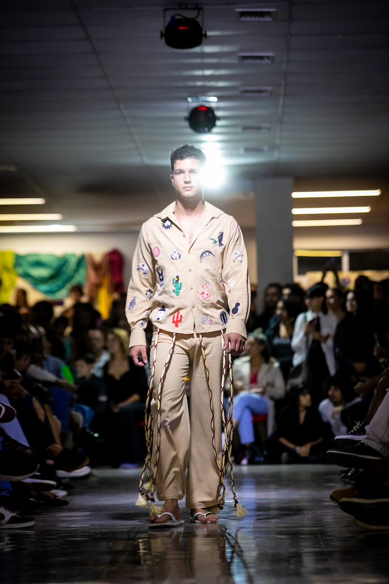 Homem caminha em passarela vestindo conjunto bege de camisa e calça com detalhes coloridos