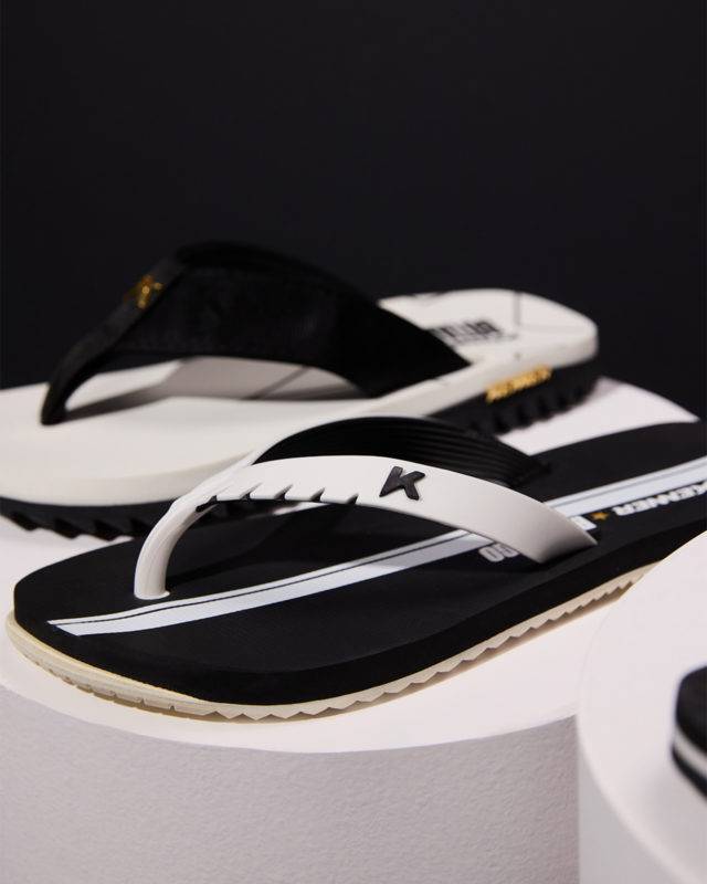 A imagem mostra duas sandálias dispostas sob uma base branca com o fundo em preto. O calçados, trabalham as mesmas cores e trazem detalhes em dourado.