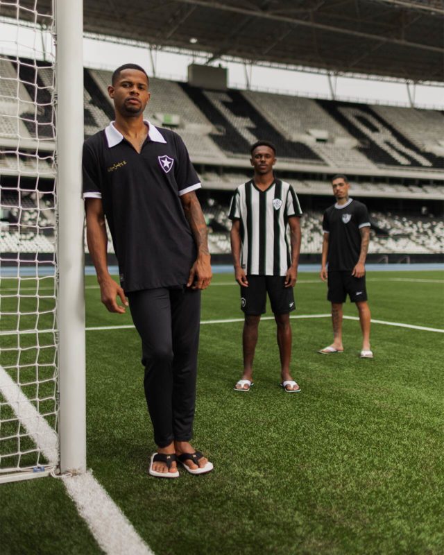 Três homens posam para a foto. Dentro de um campo de futebol, posicionados próximo ao gol, eles olham para a foto enquanto vestem trajes seguindo as cores do time Botafogo, preto e branco.