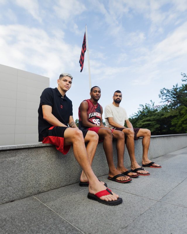 Três homens sentados posam para foto. Vestindo roupas nas cores do Flamengo, preto, vermelho e branco, eles calçam as sandálias clube em frente a brandeira do rubro negro.