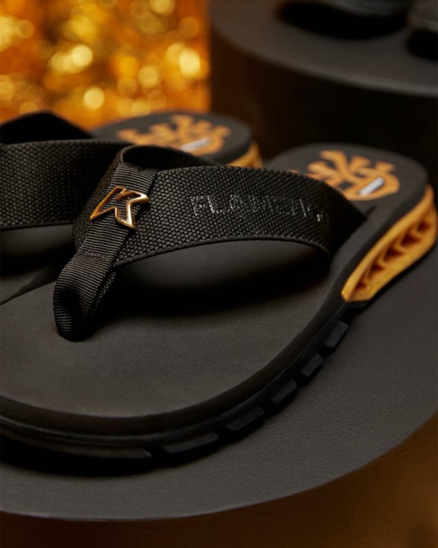 A imagem mostra uma sandália preta com detalhes dourado. Na região do calcanhar o modelo carrega a logo CRF, também em dourado.