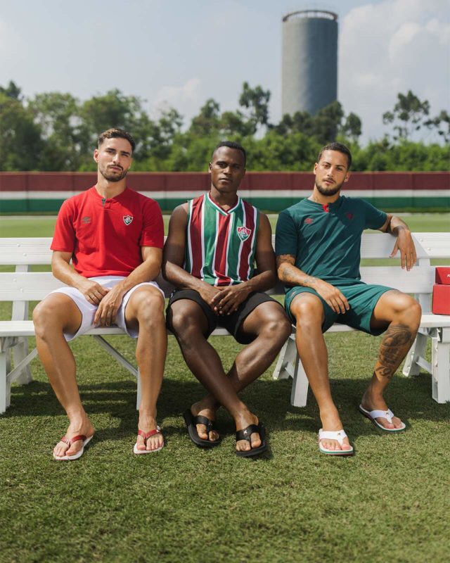 Três homens se sentados em um banco posam para a foto. Com suas roupas variando nas cores do time, vermelho, verde e branco, os rapases posam calçando as sandálias.