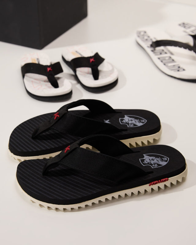 A imagem mostra três sandálias dispostas sobre uma base. Em foco, um sandália preta com detalhes em vermelho e branco.