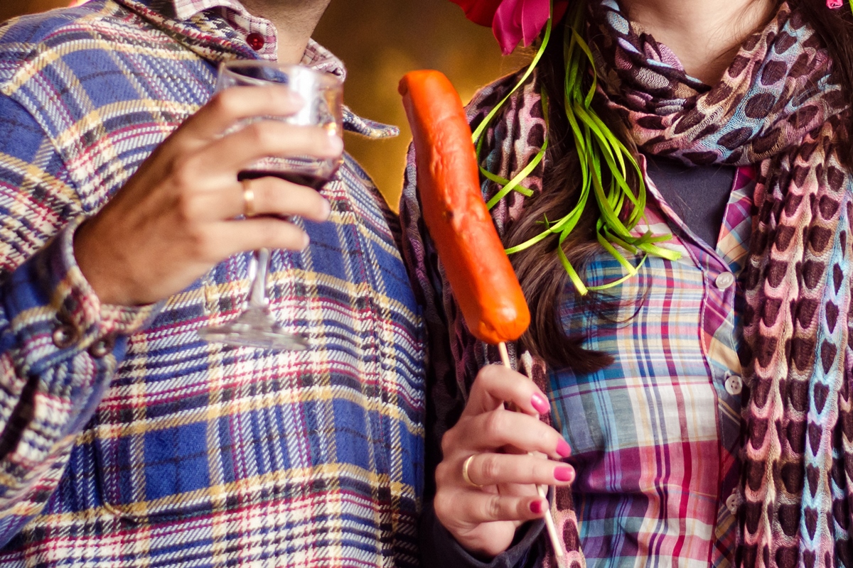 Homem e mulher segurando taça de bebida e salsichão e usando traje de festa junina - Metrópoles