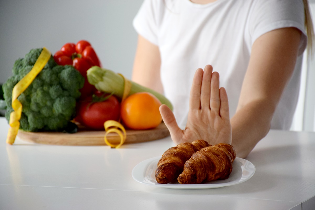 Foto colorida - Mulher com um prato repleto de fritas e legumes e com a mão em frente a outro prato com um croissant