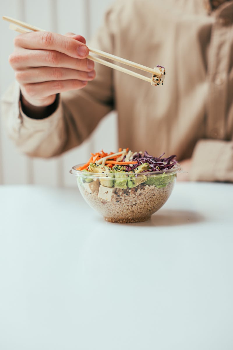 Mãos segurando hashi para pegar alimentos em um bowl transparente - Metrópoles