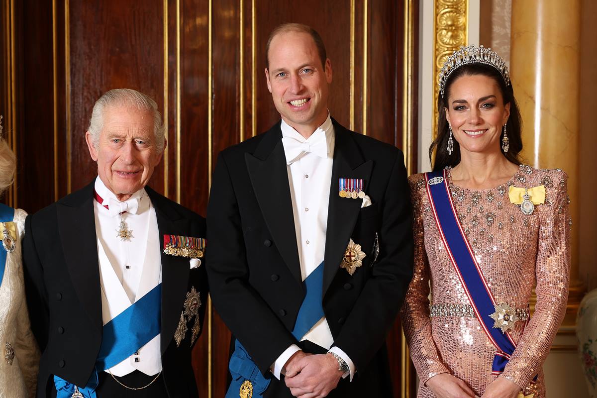 Foto colorida de dois homens com fraque uma faixa azul. Eles estão ao lado de mulher com tiara e roupa com brilhos - Metrópoles