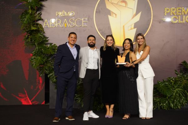 Foto colorida de cinco pessoas segurando um troféu em frente a um painel escrito Prêmio Abrasce - metrópoles 