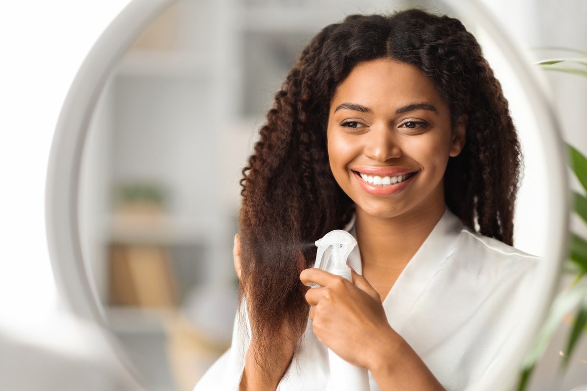 Foto de mulher se olhando no espelho enquanto aplica um produto em spray no cabelo - Metrópoles