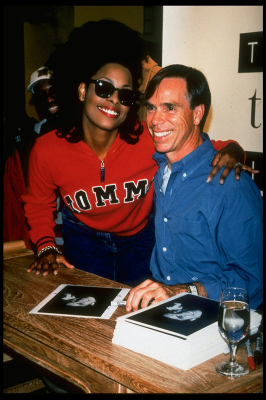 Homem e mulher sorriem em foto. Apoiada no homem, a moça de óculos escuro olha para a câmera vestindo um casaco vermelho. O homem olha para outra direção e veste uma camisa jeans azul