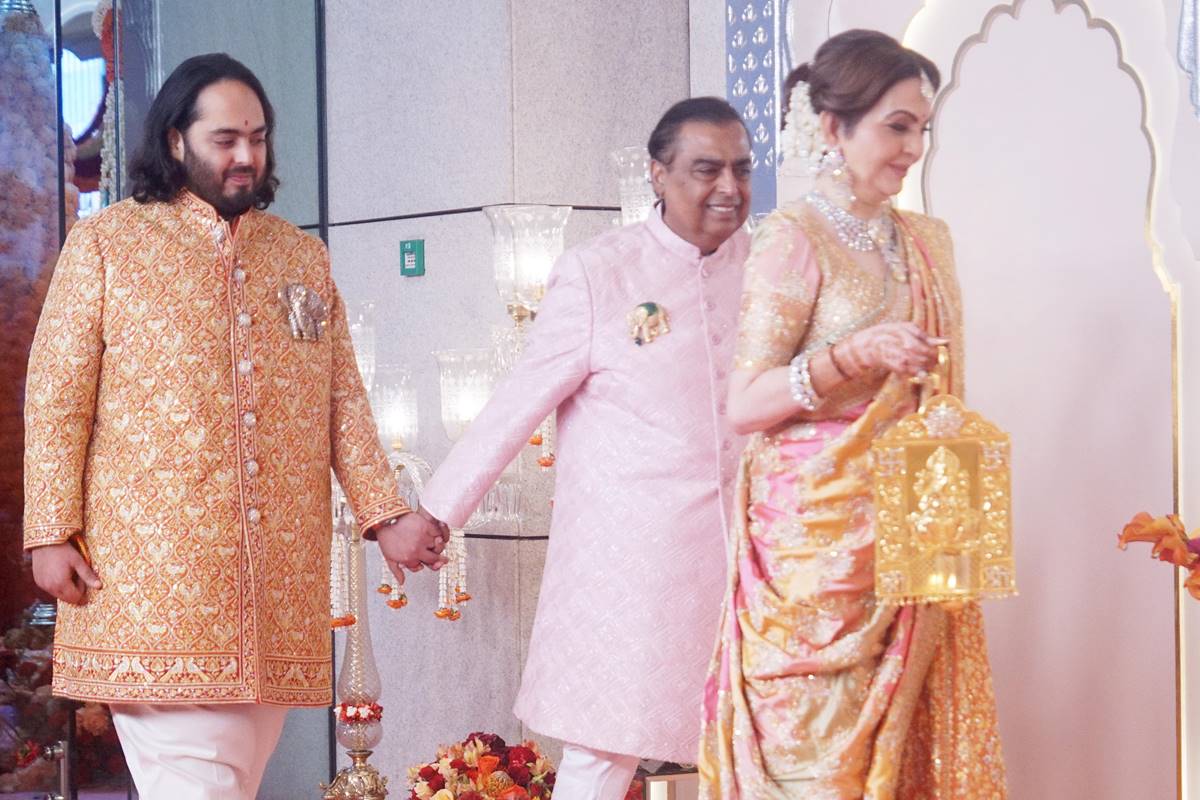 Foto colorida de homens de mãos dadas e uma mulher caminha à frente. Eles estão com trajes indianos - Metrópoles