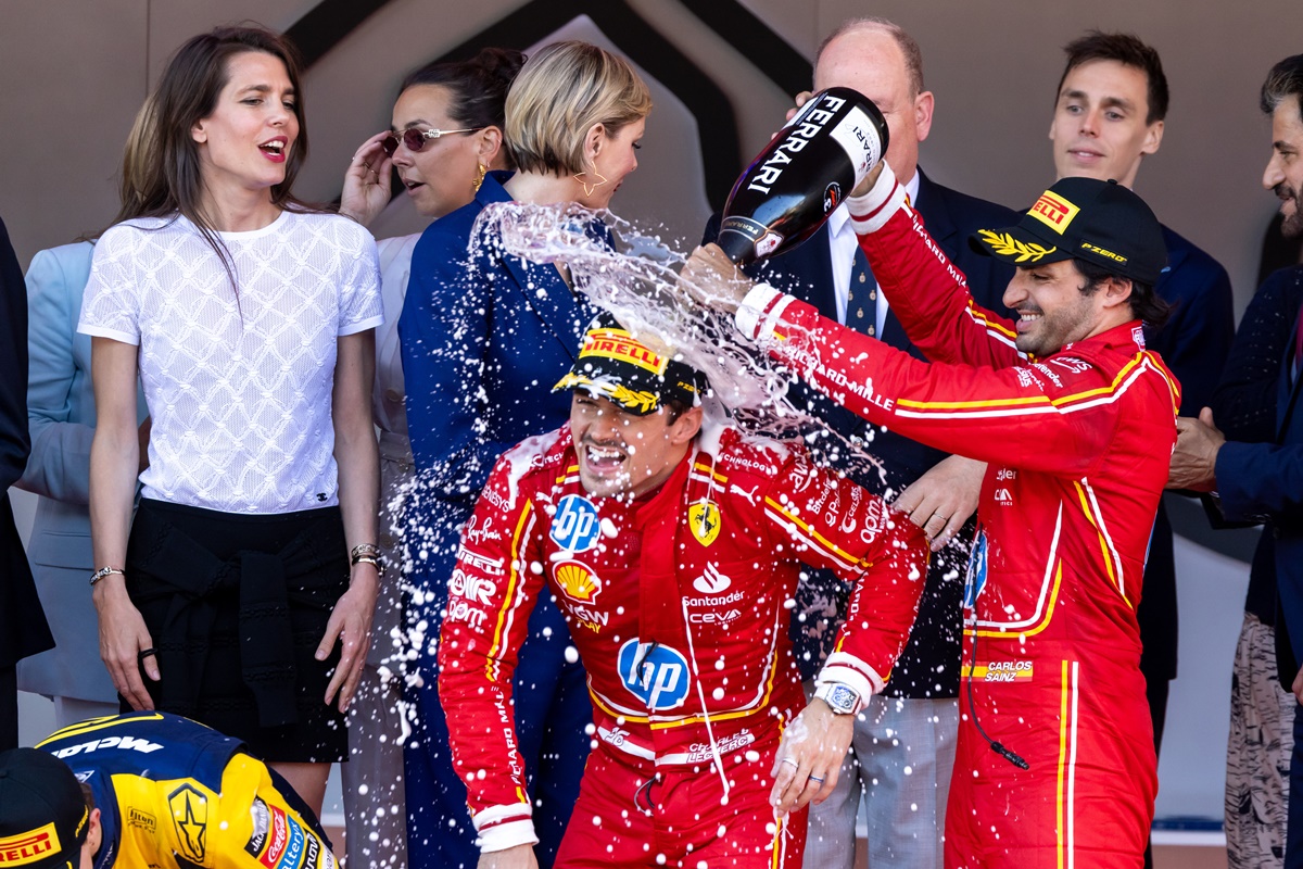 A imagem mostra um grupo de pessoas celebrando. Dois homens de macacão vermelhos com detalhes brancos e amarelos tomam o foco. Posicionado a direita, um vira uma garrafa de espumante no homem à esquera da imagem. Ambos estão rodeados de pessoas que observam o momento.