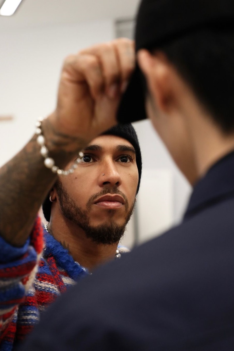 A imagem mostra um homem com um casaco vermelho, azul e branco erguendo seu braço com uma pulseira para arrumar o boné usado pelo homem em sua frente.