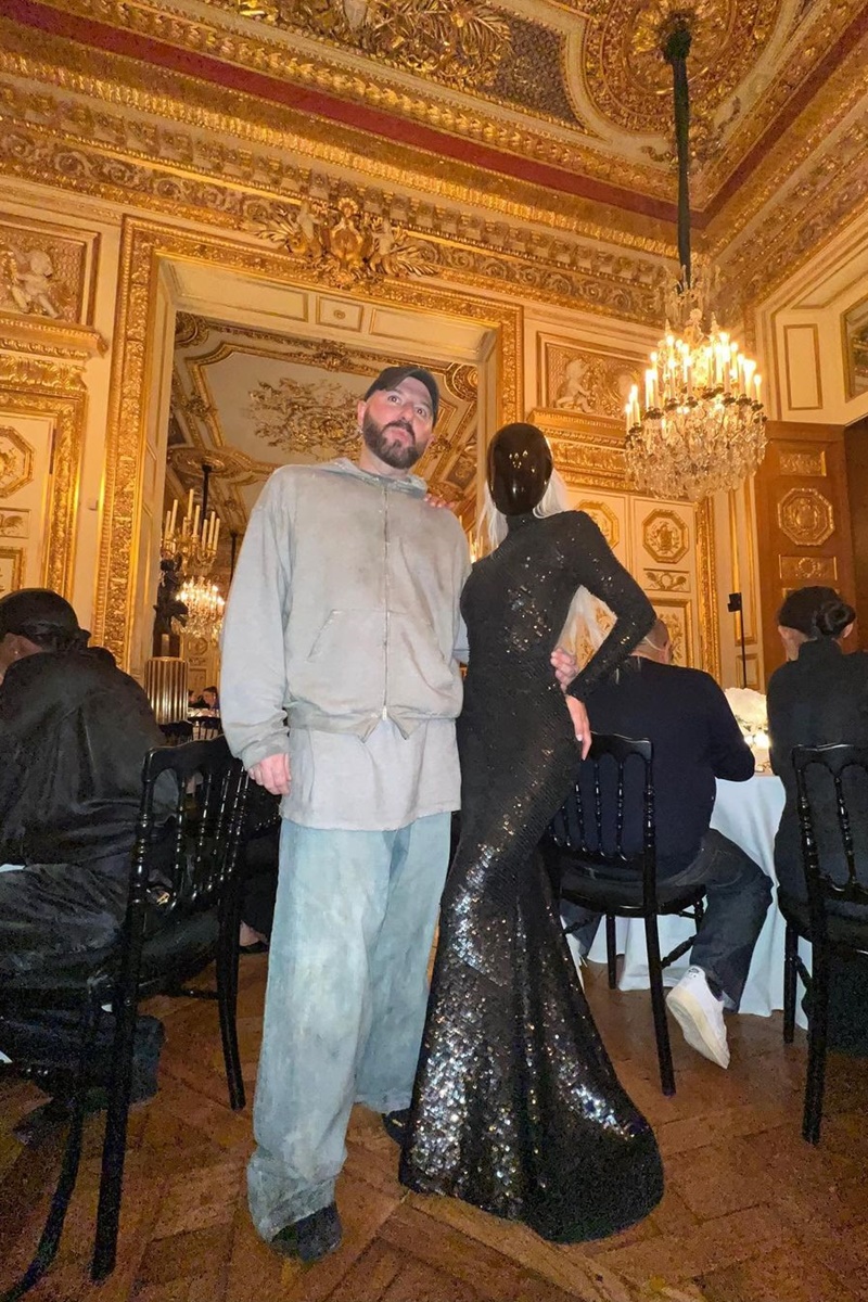 Homem e mulher posam para a foto. Em uma sala com relevos dourados, o homem veste um casaco sobrepondo sua camiseta cinza, combinando com um calça azul clara, enquanto a mulher veste um vestido preto com brilhos, cobrindo o rosto com uma capacete preto.