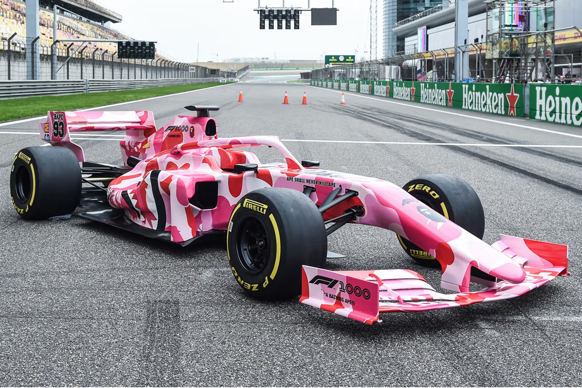 A imagem mostra um carro de fórmula 1 estacionado para ser fotografado em um autódromo. O veículo está colorido na cor rosa com uma padronagem camuflada.