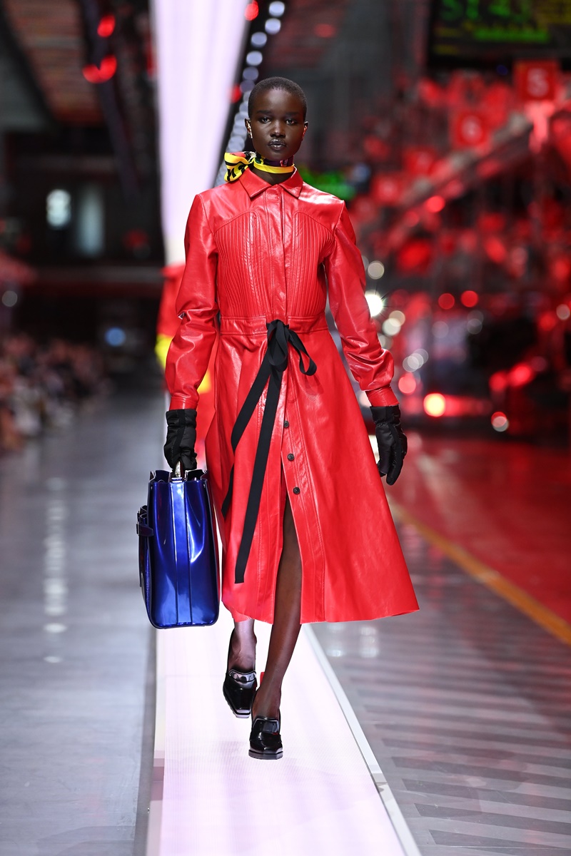 Mulher posa para a foto. Vestindo um casaco sobretudo na cor vermelha, a mulher desfile enquanto carrega uma bolsa azul em sua mão direita, coberta por uma luva preta, a qual se repete em sua outra mão. No pescoço, está amarrada uma fita amarela, na cintura uma faixa preta forma um laço e seus calçados preto, deixam parte do pé exposto.