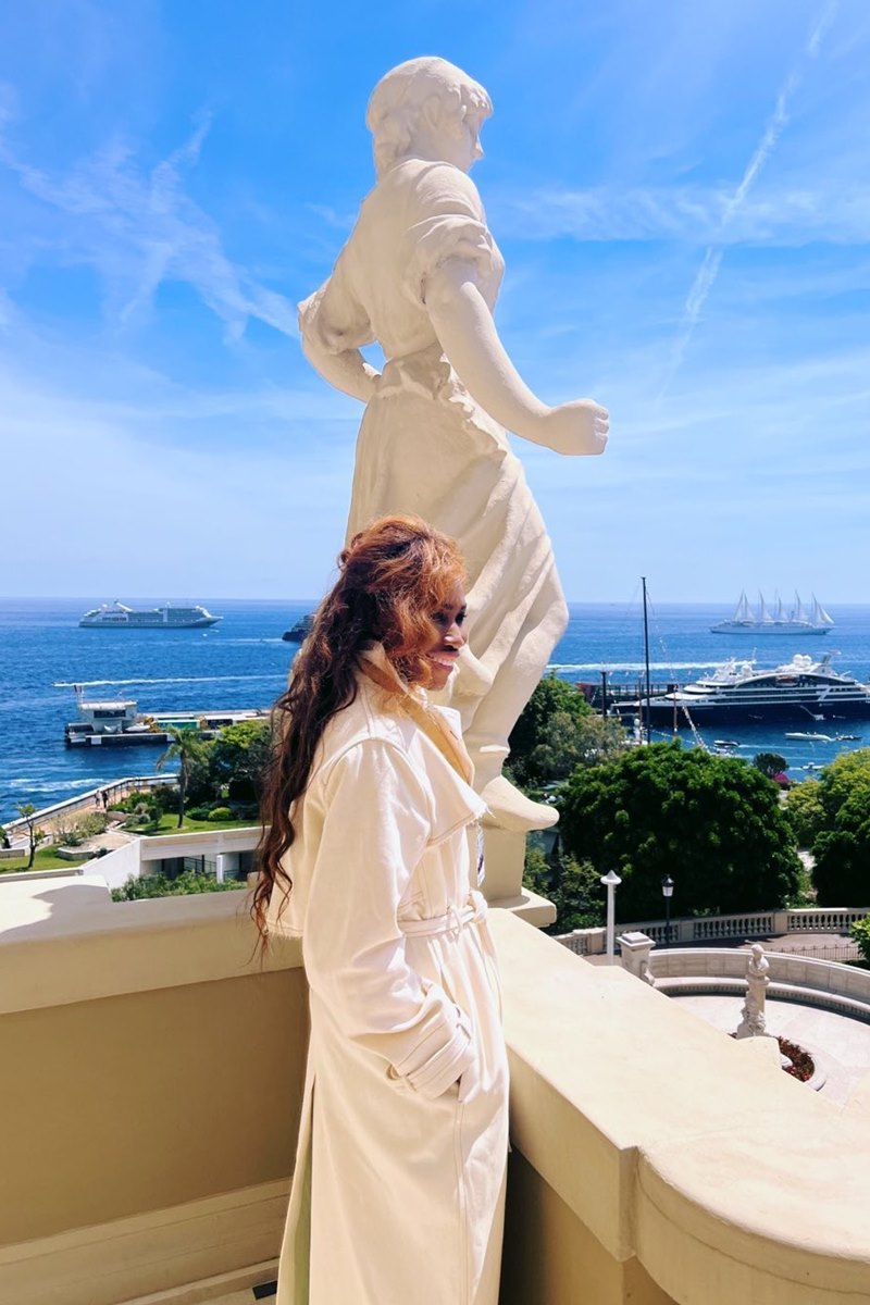 Foto colorida de mulher com casaco longo branco e perto de uma estátua - Metrópoles