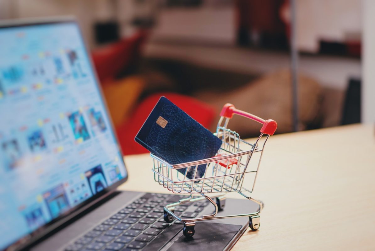 Foto colorida de carrinho de compras em miniatura com cartão de crédito dentro. Por trás, um computador - Metrópoles