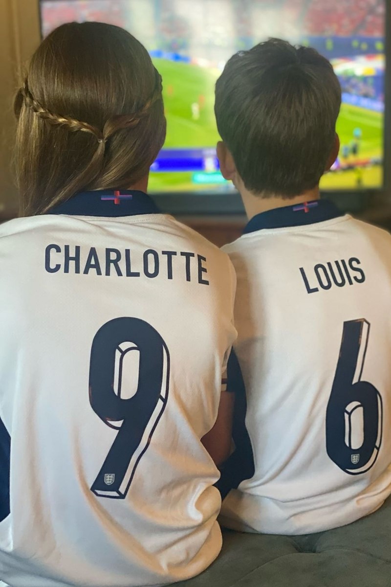 Foto colorida de menina e menino, de costas, com a blusa da seleção da Inglaterra. Eles assistem a um jogo de futebol na televisão - Metrópoles