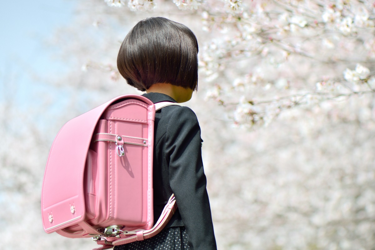 Garota posa para a foto. Em frente a uma árvore com flores rosas, a menina suegre de costas para a imagem, vestindo um casaco preto e carregando sua mochila