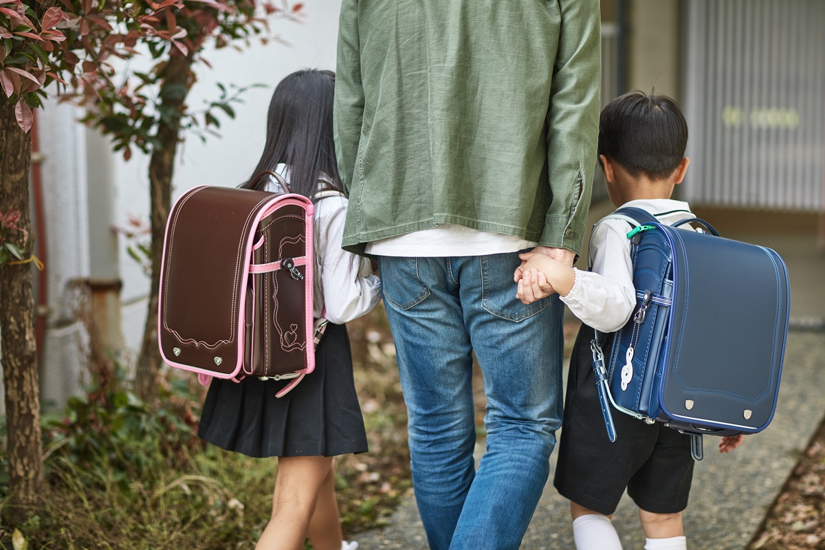 Três pessoas aparecem na imagem. De costas, duas crianças estão de mão dadas à um homem no meio. A direita, um garoto carrega uma bolsa azul e a esquerda, uma menina leva uma bolsa marrom com bordas rosas.