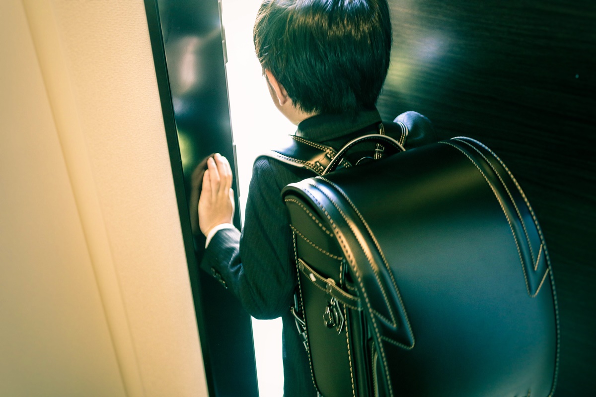 Rapaz posa para a foto. Abrindo uma porta, o jovem menino apoia sua mão na lateral enquanto abre a porta. Vestindo um terno preto listrado, ele carrega sua randoseru de couro preto nas costas.