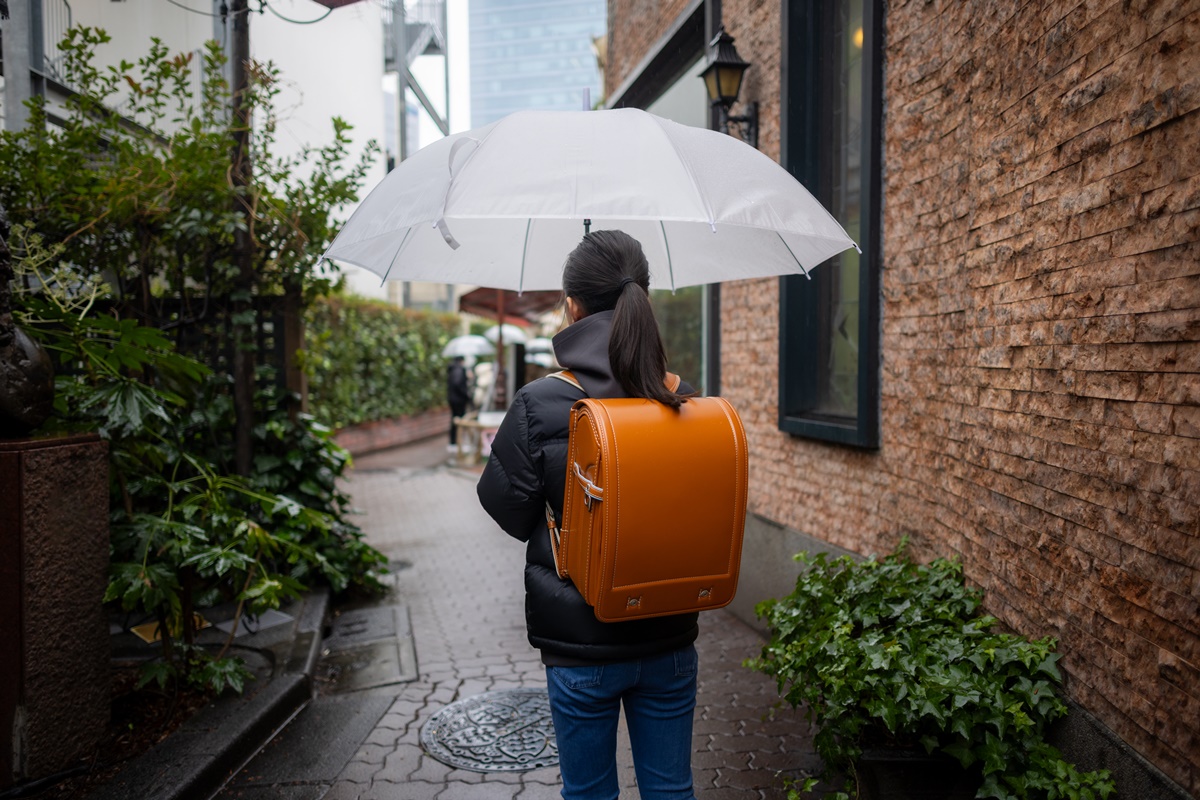 Mulher posa para a foto. De costas, ela exibe sua mochila em tom caramelho enquanto se protege da chuva com seu guarda-chuva branco. A mulher veste um casaco preto e calça jeans azul enquanto caminha.