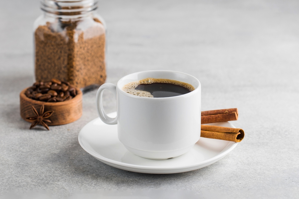 Foto de uma xícara branca com café preto e paus de canela ao lado. fundo branco - Metrópoles