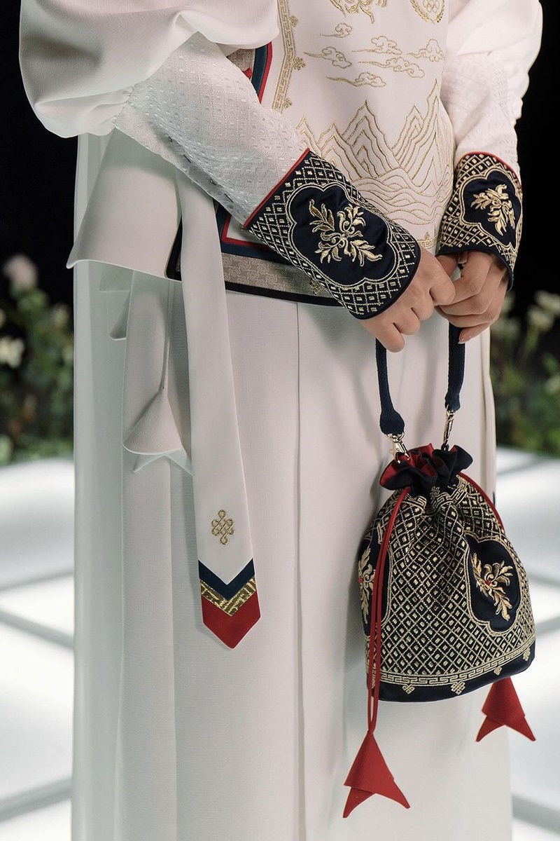 A imagem exibe uma mão segurando uma bolsa azul com bordados dourados e detalhes vermelhos. A personagem segurando a bolsa veste um roupa branca com o pulso da manga na cor azul com bordado dourado.