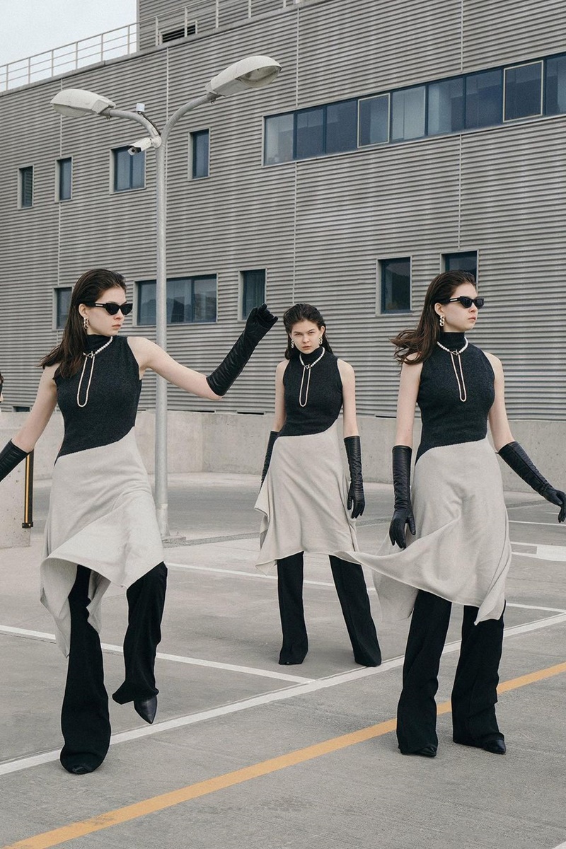 A imagem mostram uma mulher como foco ao centro da foto. Acompanhada de clones no mesmo cenário, ela veste uma camisa regata com uma saia bege, calça e luvas pretas e um colar. 