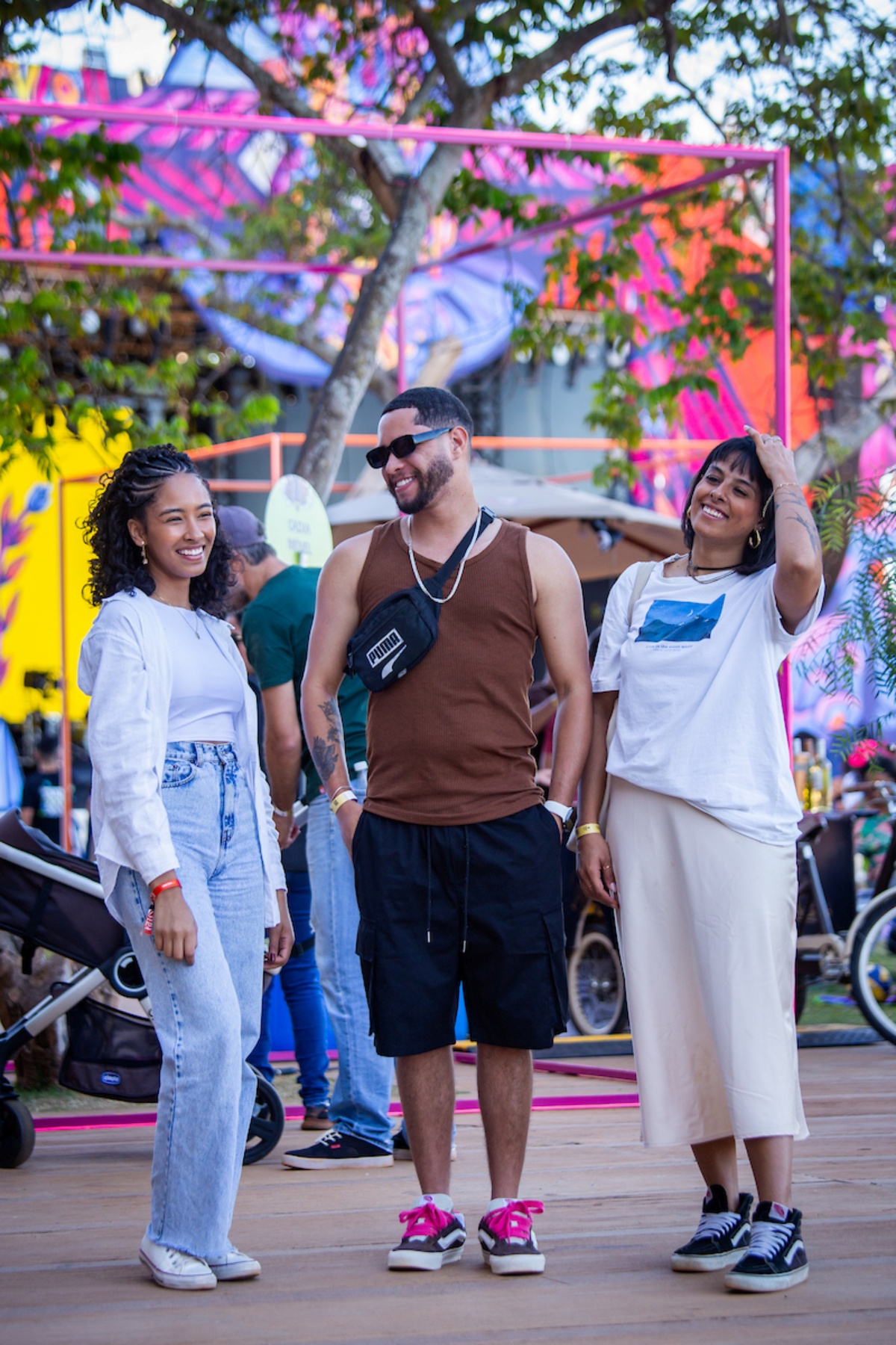 Três pessoas posam para a foto. Ao meio, um rapaz veste uma camiseta regata com uma bolsa preta curzando os ombros. A esquerda, a jovem veste uma camiseta e casaco branco, combinados com uma calça azul e a direita, outra mulher veste uma camiseta branca com uma saia langa na cor palha