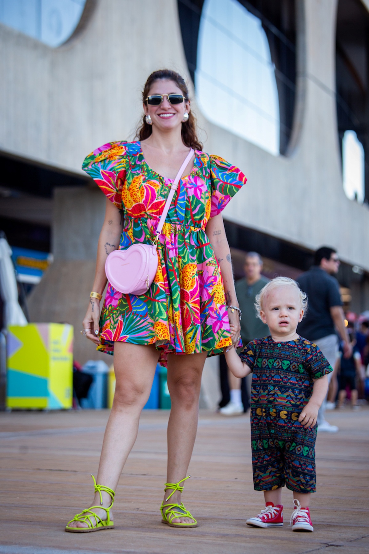 Mulher e bebê posam para a foto. Com seu vestido florido nas cores rosa, amarelo, verde, vermelho, laranja e azul, a mulher usa seus óculos escuros e bolsa rosa enquando está de mãos dadas com um bebê vestindo uma roupa preta estampada nas cores amarela, vermelha, azul e verde.