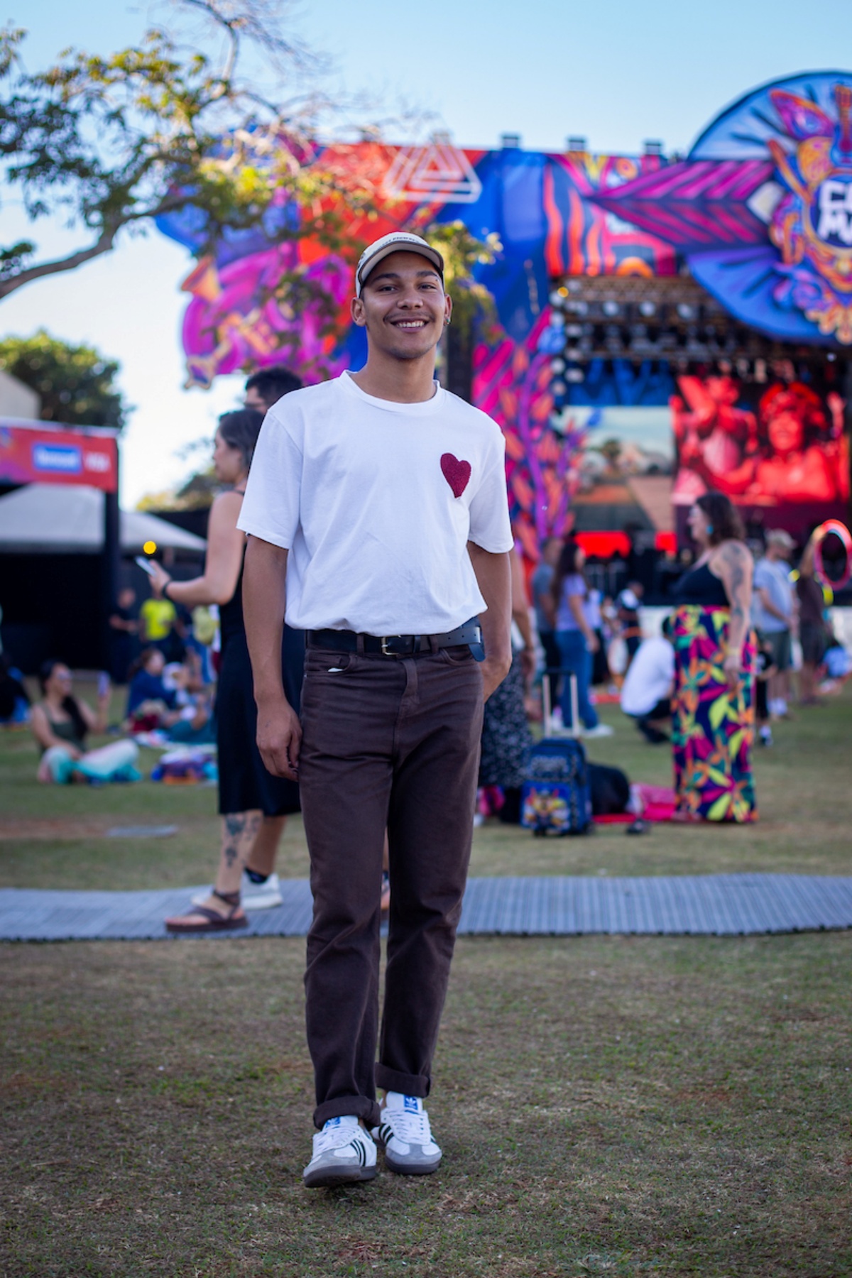 Rapaz posa para a foto. Olhando para a câmera, ele usa um boné branco em sua cabeça junto de uma camisa branca com um coração em seu peito esquerdo. Simulando um passo, ele veste também uma calça marrom e exibe seu calçado branco com detalhes em preto, cinza e marrom