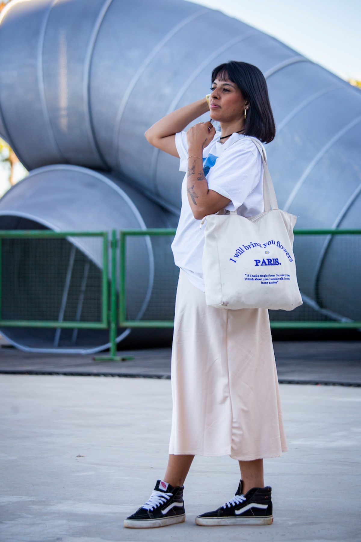 Mulher posa para a foto. Virada para a esquerda, ela usa uma camiseta branca com estampa azul, combinada com uma bolga grande com letras em azul e uma saia em tom palha