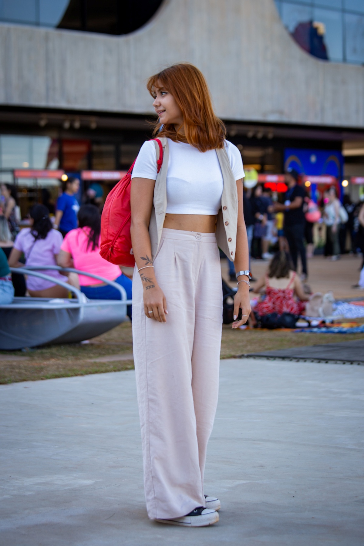 Menina posa para a foto. A moça olha para o seu lado direito enquanto carrega uma mochila vermelha. Vestindo uma camiseta branca, ela completa seu visual com um conjunto de colete e calça em tom palha.