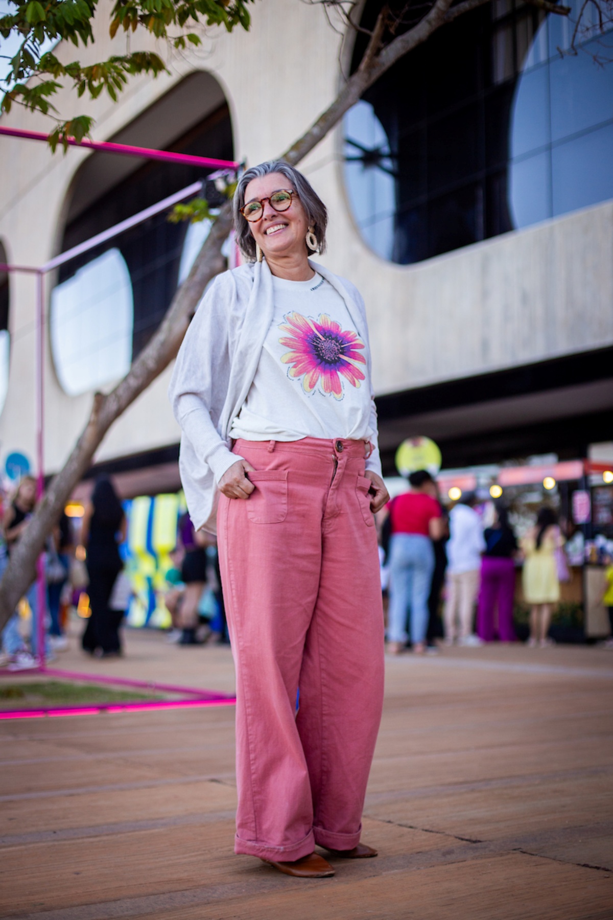 Mulher posa para a foto. Olhando para o horizonte, ela veste uma camiseta branca estampada com uma flor rosa junto de um casaco cinza e uma calça rosa.