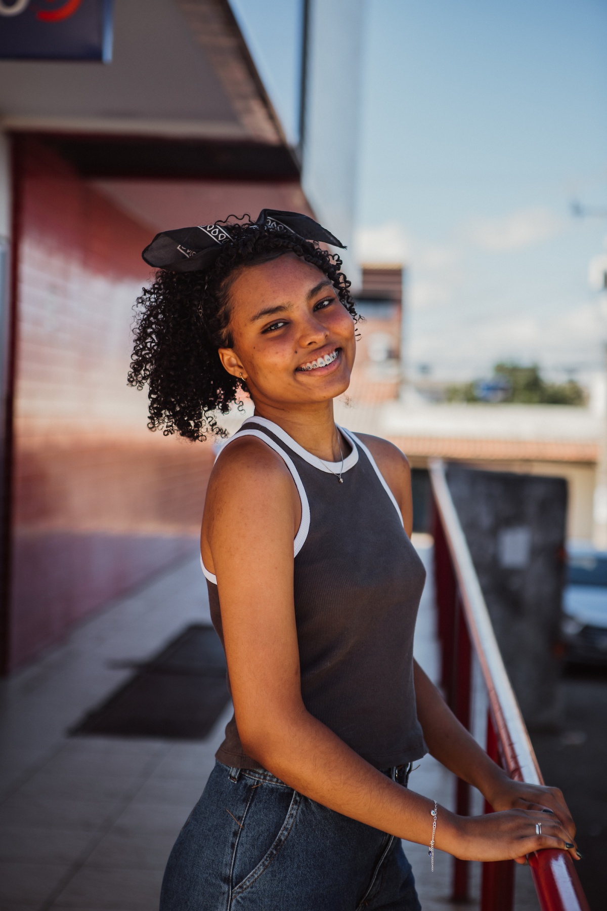 Moça posa para a foto. Olhando para a câmera, a jovem apoia suas duas mão em um corrimão à direita. Na imagem ela veste uma camiseta regata marrom com bordas brancas e um laço preto na cabeça.