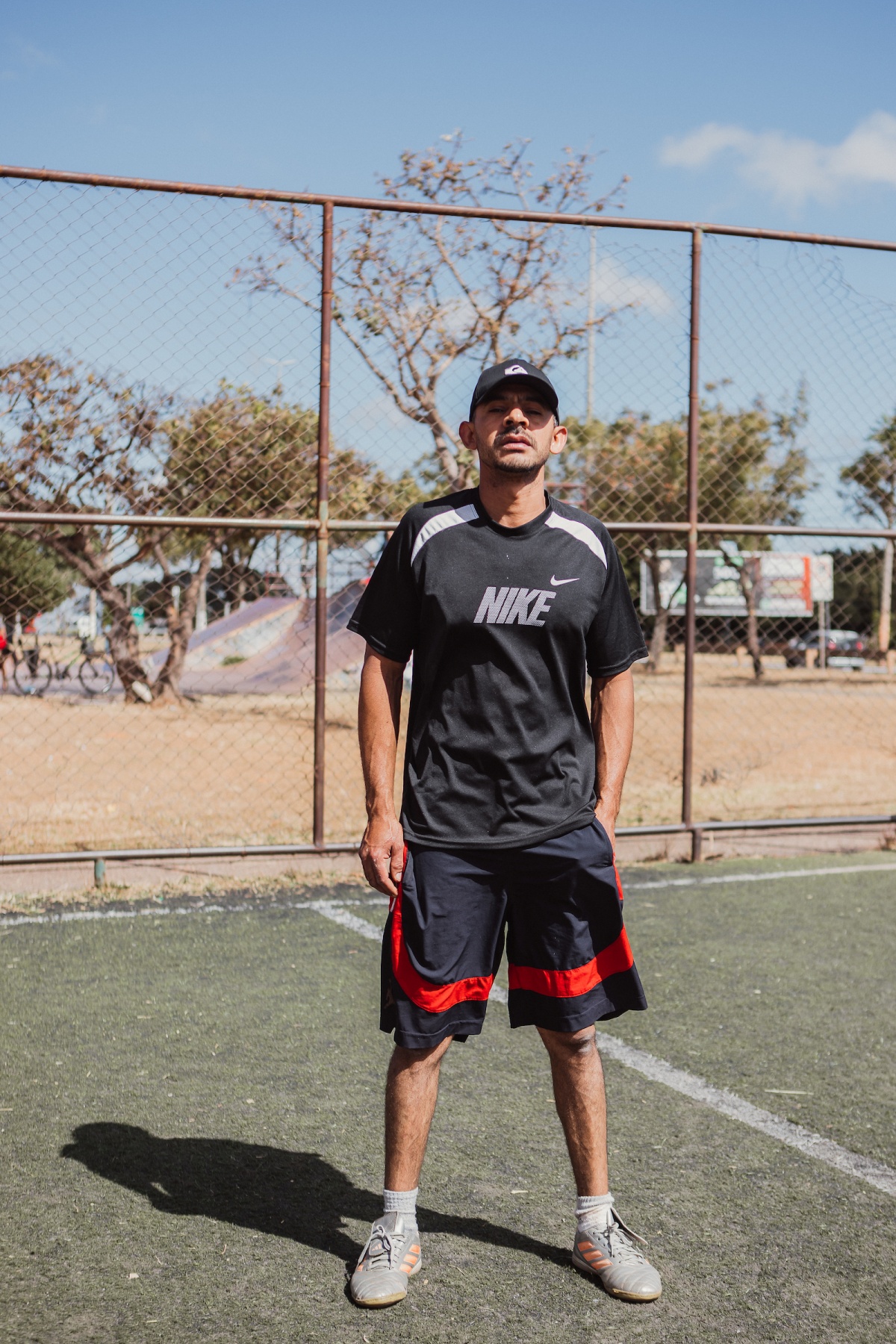 Homem posa para a foto. Olhando para a câmera, ele veste uma camiseta esportiva preta com detalhes brancos e uma bermuda preta com detalhes vermelhos. Na cabeça, o homem também usa um boné de aba torta