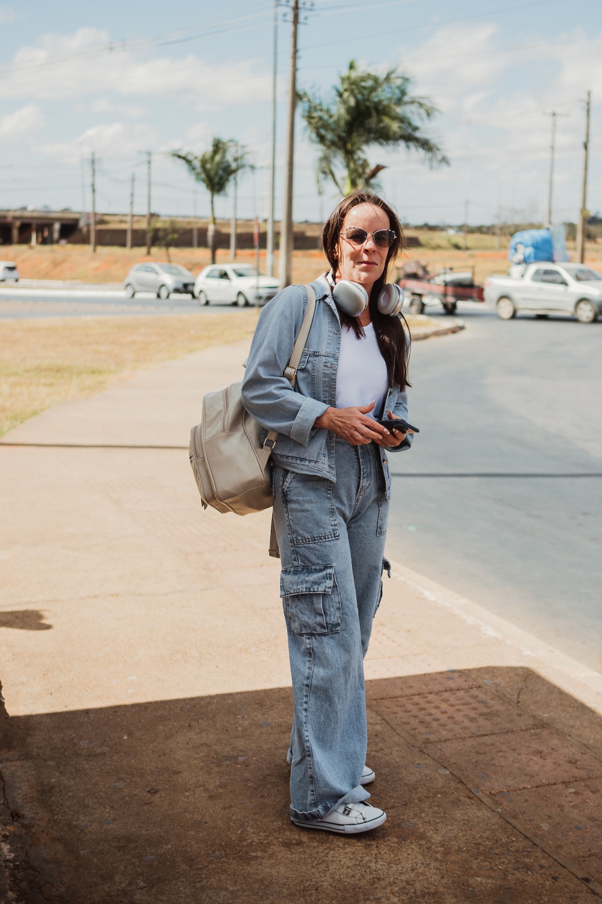 Mulher posa para a foto. Com um conjunto de calça e camisa jeans, ela carrega uma mochila na cor branco creme nas costas e um fone da mesma cor em seu pescoço.